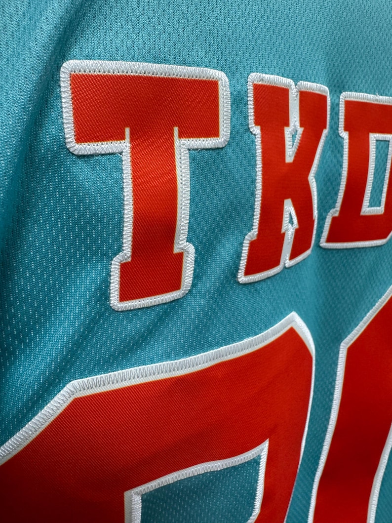 May include: Close-up of a turquoise jersey with red lettering and numbers. The letters and numbers are outlined in white stitching. The jersey has a mesh texture.