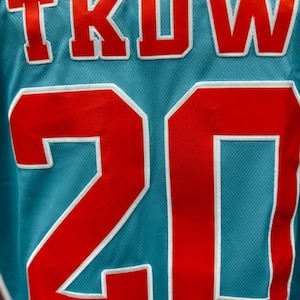 May include: Close-up of a teal jersey with the letters "TKOW" in red with white trim at the top and the number "20" in red with white trim at the bottom. The jersey has a mesh texture.