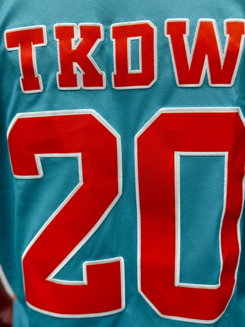 May include: Close-up of a teal jersey with the letters "TKOW" in red with white trim at the top and the number "20" in red with white trim at the bottom. The jersey has a mesh texture.