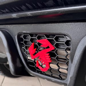 Peut inclure: Un emblème de scorpion rouge est fixé à une calandre noire en nid d'abeille sur une voiture.