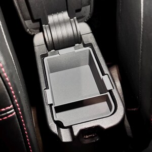 May include: Black leather car center console with a gray plastic storage compartment. The compartment has a lid that opens to reveal a small storage space.