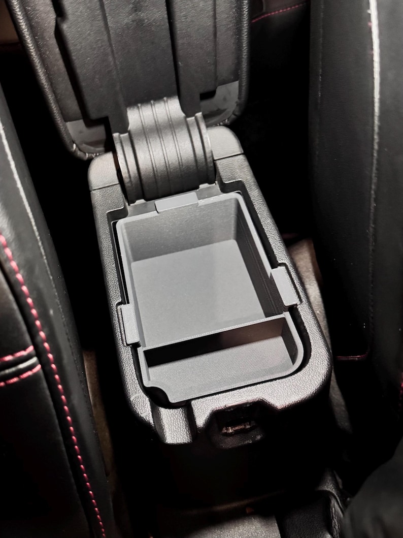 May include: Black leather car center console with a gray plastic storage compartment. The compartment has a lid that opens to reveal a small storage space.