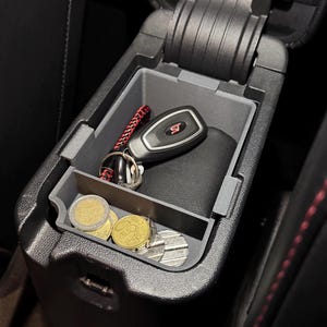 May include: A black car center console with a gray organizer insert. The organizer has a compartment for a key fob and a smaller compartment for coins. The key fob has a red "ST" logo. There are several gold coins in the coin compartment.