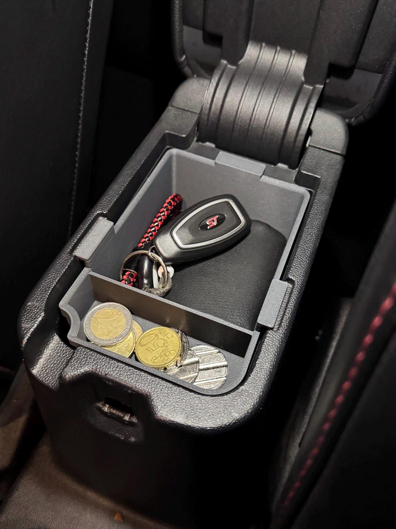 May include: A black car center console with a gray organizer insert. The organizer has a compartment for a key fob and a smaller compartment for coins. The key fob has a red "ST" logo. There are several gold coins in the coin compartment.