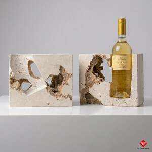 Può includere: Due portabottiglie in pietra strutturata bianco sporco con aperture irregolari ed erose. Un supporto contiene una bottiglia di vino con un'etichetta dorata. L'etichetta recita "ANSELMI 2015 SAN VINCENZO".