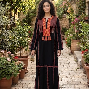 Puede incluir: Vestido largo negro con bordados rojos y naranjas. El vestido tiene un escote en V y mangas largas. El bordado se concentra alrededor del cuello, las mangas y el dobladillo. El vestido se usa al aire libre en un jardín.