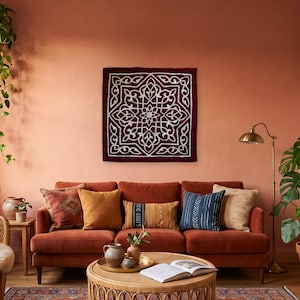 Puede incluir: Un tapiz cuadrado con una base de tela color burdeos intenso y un patrón geométrico blanco, intrincado y simétrico. El diseño consta de líneas entrelazadas y motivos florales centrados dentro de un marco de borde cuadrado.