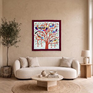 May include: A square wall hanging featuring a central tree with a thick red trunk and branches. The tree is adorned with stylized birds in shades of blue, yellow, and purple, surrounded by green pine-like needles and small red circular accents.