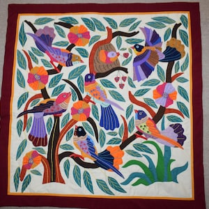 Puede incluir: Un tapiz textil cuadrado con un centro color crema y un borde rojo oscuro. El diseño muestra pájaros coloridos, flores naranjas y moradas, y hojas verdes bordadas sobre ramas. Se puede ver un nido de pájaro con un ave pequeña.