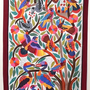 May include: A rectangular textile wall hanging featuring intricate embroidery of numerous colorful birds perched on branches amidst green leaves and pink flowers. The design is bordered by a thick dark red frame on a white background.