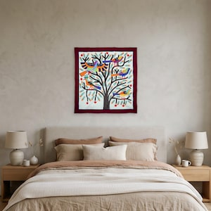 May include: A square textile wall hanging featuring a central tree with black branches and trunk on a white background. The tree is decorated with colorful birds in shades of blue, orange, and yellow, along with small red circular accents.