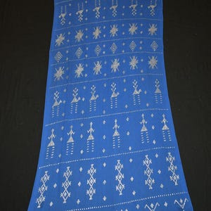 Puede incluir: Textil azul largo y rectangular con un patrón repetitivo de figuras geométricas y humanas estilizadas plateadas. La tela tiene una textura suave y probablemente está hecha de algodón o un material similar. El diseño es simétrico y cubre toda la superficie.