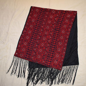 May include: A rectangular scarf with a red and black floral pattern and black fringe. The scarf has a black backing and a detailed, repeating design in red and white on a black background.