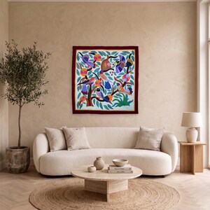 May include: A square wall hanging featuring a vibrant, multi-colored illustration of various birds perched on stylized tree branches with green leaves, all enclosed within a solid dark red border on a light blue background.