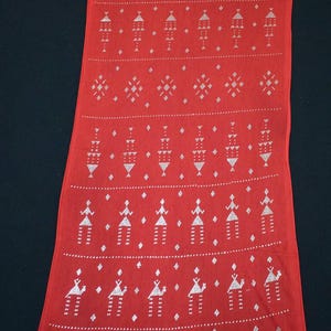 May include: A long, rectangular red textile with a repeating white geometric pattern. The design includes diamond shapes, stylized figures, and arrow-like motifs. The fabric appears to be lightweight and suitable for a scarf or shawl.