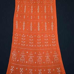 May include: An orange textile with a repeating pattern of white geometric shapes. The design includes diamond, arrow, and triangle motifs, creating a symmetrical and detailed visual texture. The fabric appears to be a lightweight material.