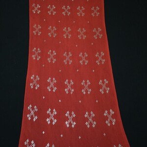 May include: A long, rectangular red textile with a repeating pattern of silver cross-like designs. The top and bottom edges feature decorative borders with small silver details. The fabric is draped against a black background.