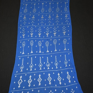 May include: A long, rectangular blue scarf with a repeating silver geometric pattern. The pattern includes diamonds, triangles, and stylized tree-like shapes. The scarf is displayed against a dark background.