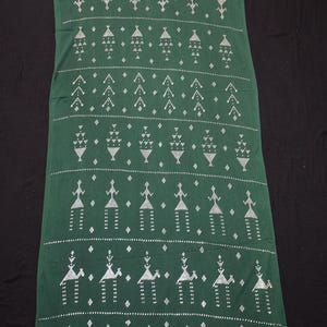 Puede incluir: Un textil largo y rectangular verde con un patrón geométrico blanco repetitivo. El patrón incluye árboles estilizados, formas de diamante y figuras humanas. El textil es probablemente una pieza decorativa o una bufanda.