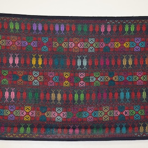 May include: A rectangular textile with a dark background and intricate, colorful embroidery. The design features rows of repeating geometric and floral patterns in shades of red, green, yellow, purple, and blue. The edges are finished with a dark border.