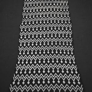 Puede incluir: Un vestido con estampado en blanco y negro con un diseño geométrico. El vestido presenta un patrón repetitivo de zigzag y diamantes en blanco sobre un fondo negro. El vestido se muestra sobre una superficie negra.
