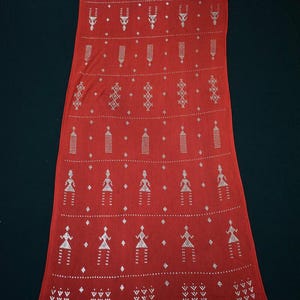 May include: A long, rectangular red textile with a repeating pattern of silver-colored geometric and stylized human figures. The design includes diamond shapes, triangles, and other abstract forms. The textile is likely a scarf or shawl.