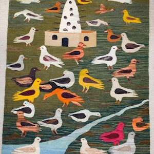 May include: Hand-woven rug with a green background featuring a whimsical design of birds in various colors, including white, yellow, orange, and pink. A stylized building with a tower is in the center. The rug has a fringed edge.