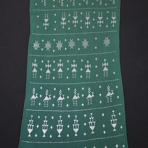 Puede incluir: Un textil largo y rectangular verde con un patrón geométrico blanco repetitivo. El diseño incluye filas de figuras estilizadas, diamantes y otras formas. El textil es probablemente una bufanda o una pieza decorativa.