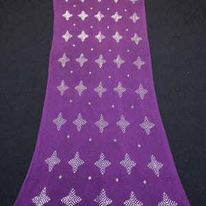 May include: A long, rectangular purple scarf with a repeating pattern of white geometric starburst designs. The scarf has a white dotted border along the bottom edge and is made of a soft fabric.