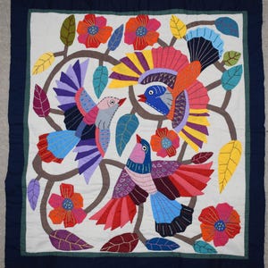 Pode incluir: Uma tapeçaria têxtil quadrada apresentando três pássaros coloridos em voo entre ramos castanhos, folhas verdes e amarelas, e flores vermelhas e cor de laranja. O design é definido contra um fundo branco com uma borda azul escura.