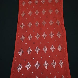 May include: A long, rectangular red scarf with a repeating silver geometric pattern. The scarf has a decorative border of silver triangles along the bottom edge and a silver dotted border along the top edge. The scarf is made of a lightweight fabric.