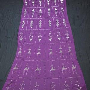 Puede incluir: Un textil largo y rectangular de color púrpura con un patrón repetitivo de formas geométricas blancas. El diseño incluye filas de figuras estilizadas y formas de diamante, creando una apariencia simétrica y decorativa.