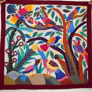 May include: A colorful textile square featuring a vibrant bird and tree design. The artwork showcases various birds with bright plumage, perched on branches with green leaves and yellow fruit. The border is a deep maroon, framing the detailed, handcrafted scene.