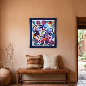 Op de afbeelding: Een vierkante textiele wandhanger met een levendig applicatieontwerp van kleurrijke vogels en bloemen op een witte achtergrond, omlijst door een donkerblauwe rand. Het kunstwerk toont ingewikkelde patronen in rood, geel, paars en groen.
