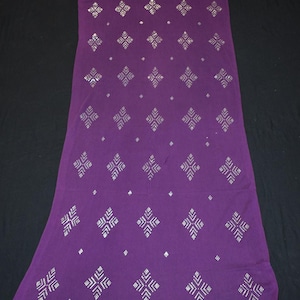Puede incluir: Un textil largo y rectangular de color púrpura con un patrón repetitivo de formas geométricas plateadas y puntos. Los bordes están acabados con un borde decorativo de triángulos y puntos. La tela está tendida sobre una superficie negra.