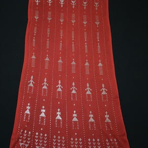 May include: A long, rectangular red scarf with a repeating pattern of silver geometric shapes and figures. The design includes rows of stylized figures and decorative elements, creating a textured appearance. The scarf is displayed against a black background.