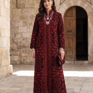 May include: A long, red and black embroidered dress with a high collar and long sleeves. The dress features intricate floral patterns and a front zipper. The person is wearing a silver necklace and bracelet, and carrying a matching clutch. The person is wearing sandals.