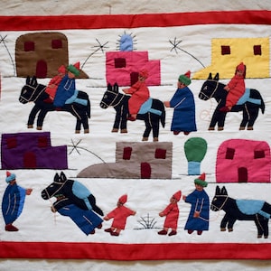 Puede incluir: Pieza de arte textil hecha a mano con figuras sobre burros, edificios coloridos y un borde rojo. Las figuras visten túnicas y sombreros de varios colores. La obra de arte es probablemente una representación de una escena bíblica.