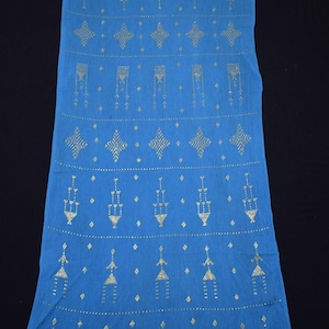 Puede incluir: Un textil azul largo y rectangular con un patrón repetido de formas geométricas doradas. El diseño incluye diamantes, triángulos y líneas verticales, creando un patrón simétrico y decorativo. La tela parece ser un material suave.