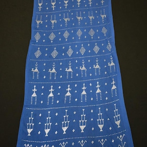 Puede incluir: Un textil azul largo y rectangular con un patrón repetitivo de formas geométricas plateadas. El diseño incluye figuras estilizadas, diamantes y motivos en forma de flecha. La tela parece suave y se drapea suavemente.