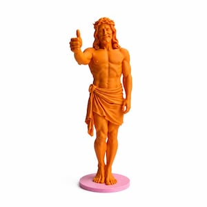 Könnte beinhalten: Orangefarbene Statue eines Mannes mit langem Haar, einer Dornenkrone und einem drapierten Tuch um die Taille, der einen Daumen hoch zeigt. Die Statue steht auf einem rosa kreisförmigen Sockel. Die Figur ist muskulös und hat einen lächelnden Gesichtsausdruck.