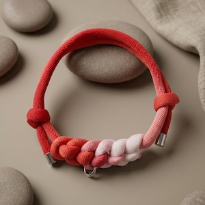 May include: A braided dog collar in shades of red, pink, and white. The collar features a gradient effect, transitioning from a deep red to a soft pink and white. It has a metal ring and metal accents. The collar is made of a thick, woven rope.