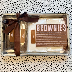 May include: A brownie baking kit presented in a silver tray, secured with a brown ribbon. The kit contains dark chocolate, milk chocolate, sugar, and instructions. The word "BROWNIES" is printed on the front of the kit.