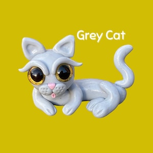 Puede incluir: Figura de gato gris claro con ojos grandes y expresivos y una nariz rosa. El gato está tumbado con la cola enroscada. El texto "Grey Cat" está encima del gato sobre un fondo amarillo.