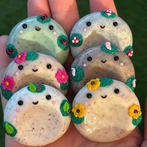 May include: Six round, smooth soap bars with smiling faces, black eyes, and pink cheeks. Each bar features small, colourful clay decorations including ladybirds, red mushrooms, pink flowers, green leaves, and yellow sunflowers attached to the surface.