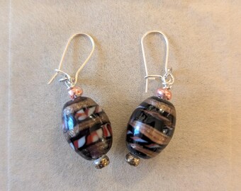 May include: A pair of dangle earrings with black, white, and gold striped glass beads. The beads are oval shaped and have a metallic sheen. The earrings have silver hooks and copper accents.