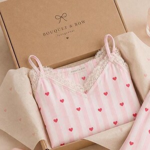May include: A pink and white striped camisole with red heart accents and lace trim, nestled in a cardboard box. The box is labeled "BOUQUET & BOW Loungewear". The camisole has thin straps and a V-neck design. The packaging includes tissue paper.