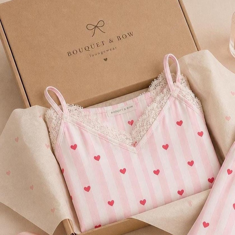 May include: A pink and white striped camisole with red heart accents and lace trim, nestled in a cardboard box. The box is labeled "BOUQUET & BOW Loungewear". The camisole has thin straps and a V-neck design. The packaging includes tissue paper.