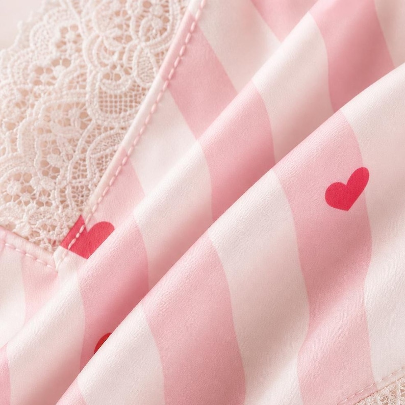 May include: Close-up of a pink and white striped fabric with red heart accents. The fabric has a silky sheen and is folded, revealing a delicate lace trim. The overall theme suggests sleepwear or lingerie.