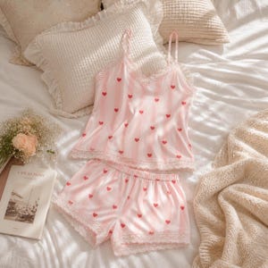 May include: A pink and white striped pajama set with red heart accents. The set includes a camisole top with lace trim and matching shorts. The pajamas are displayed on a white bed with pillows and a blanket.
